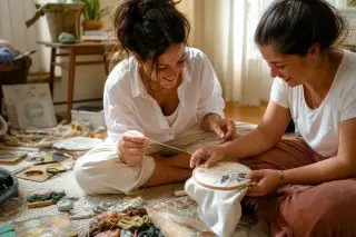 Two women exploring embroidery together on a cozy living room rug, hands busy with colorful materials, in warm natural sunlight.