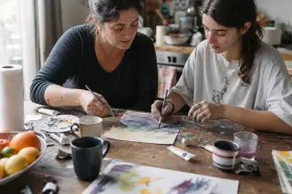 Two people of different ages painting together on a cluttered kitchen table, focused on blending colors with their hands under soft natural light.
