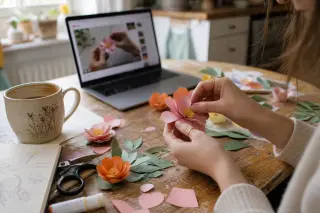 Woman's hands assembling paper flowers on a cluttered kitchen table, morning light coming through a window, laptop with a tutorial video open in the background.