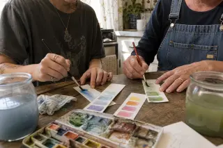 Two adults absorbed in a hands-on watercolor session at a sunlit kitchen table, surrounded by art supplies and natural light, with hands painting in focus and an inviting, relaxed atmosphere.