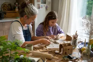 Two people of different ages shaping clay at a kitchen table filled with scattered craft materials, bathed in warm natural light, focused on their creation.