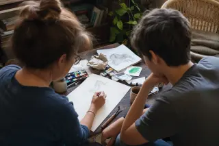 Two people of different ages working together on an improvised craft table, focused on sketching amid scattered art supplies, in a sunlit, cozy home corner.