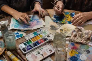 Two people’s hands at a messy kitchen table experimenting with acrylic and watercolor, natural daylight, casual and creative atmosphere.